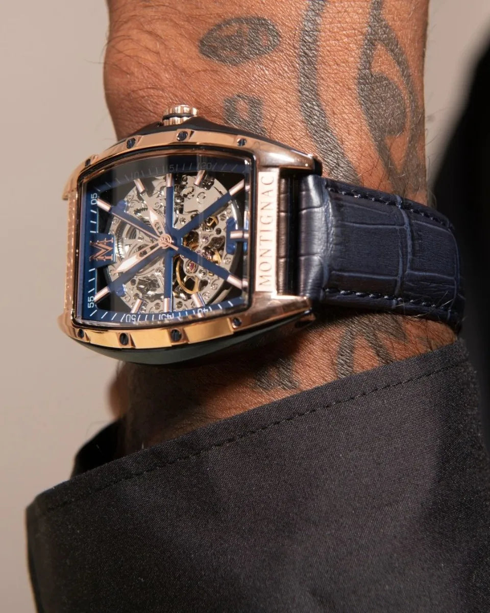 Luxury watch with blue face and black strap worn by a person with tattoos.